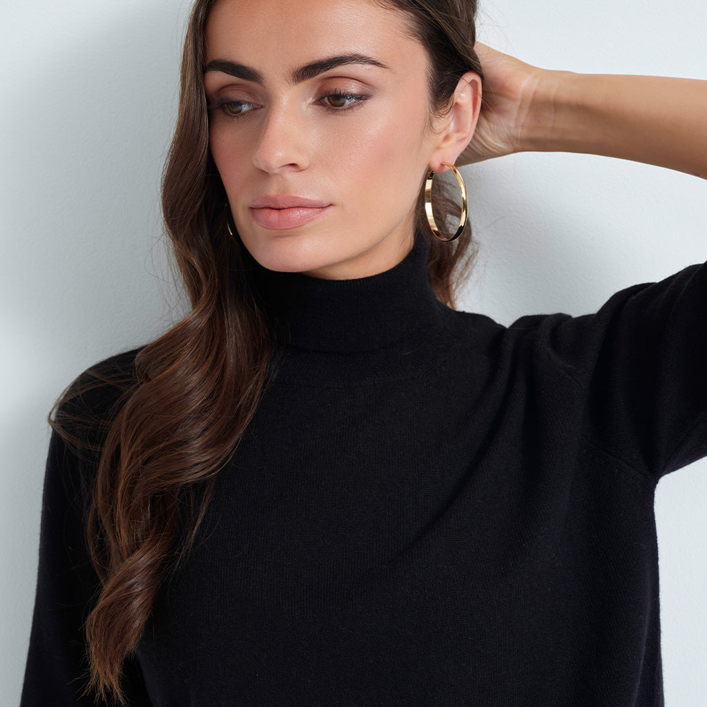 A woman wearing a black turtleneck sweater and large gold hoop earrings, shown in a close-up studio shot against a light gray background.