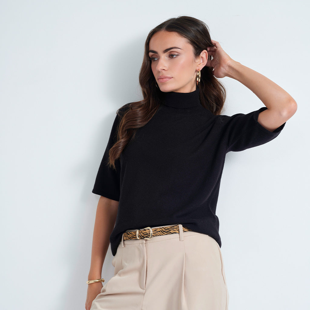 A woman wearing a black short-sleeve mock neck top and beige trousers with a patterned belt, posing against a light gray wall.
