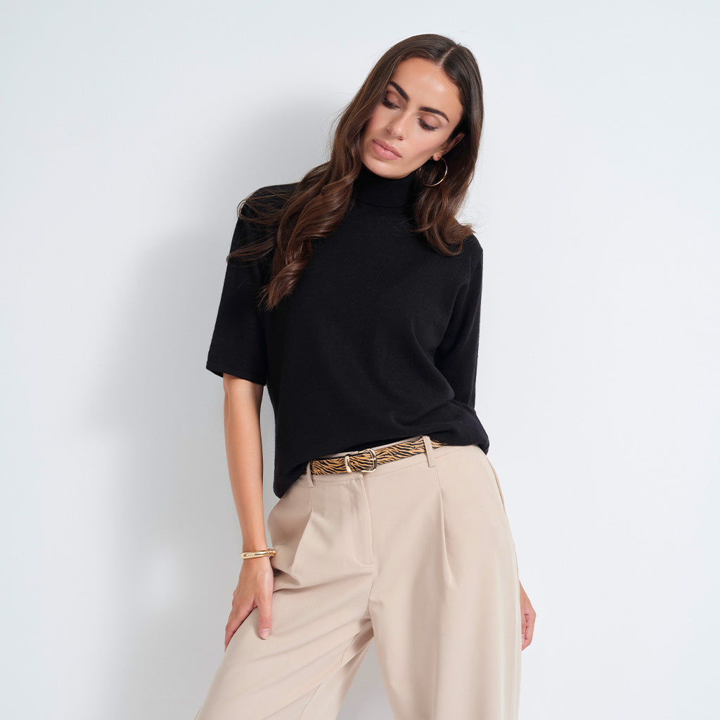 A woman posing in a black short-sleeve mock neck knit top and beige wide-leg trousers with a leopard-print belt, set against a plain light gray studio background.