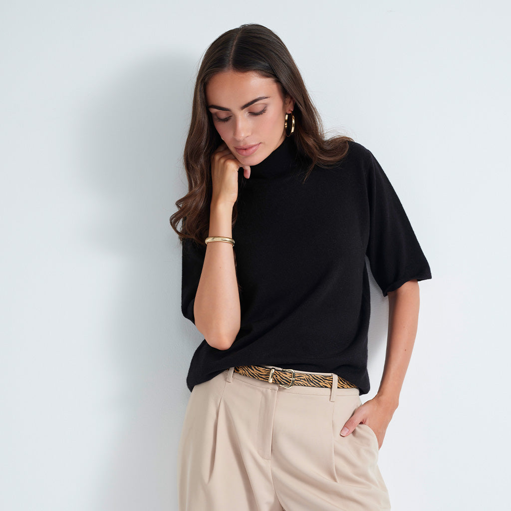A woman wearing a black short-sleeve mock neck top and beige trousers with a leopard-print belt, posing against a light gray wall.