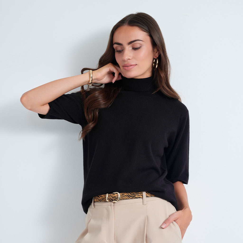 A woman wearing a black short-sleeve mock neck top and beige trousers with a patterned belt, posing against a light gray wall.