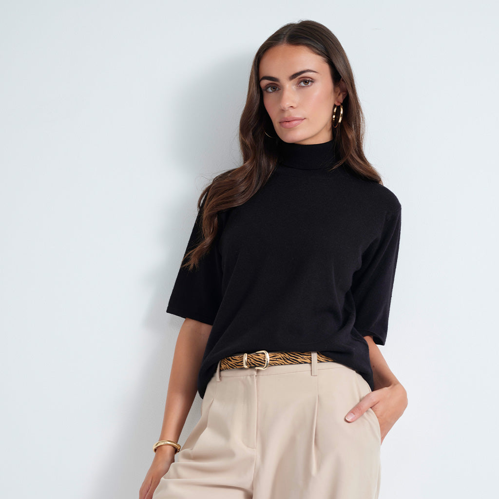 A woman modeling a black short-sleeve mock neck top paired with beige trousers and a leopard-print belt, standing against a light gray studio background.