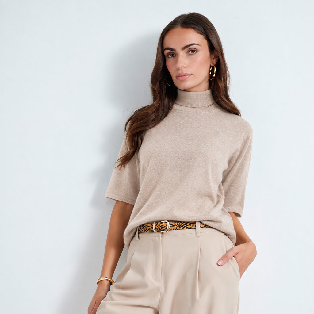 A woman modeling a beige short-sleeve mock neck knit top with light beige trousers and a patterned belt, standing against a light blue-gray studio background.