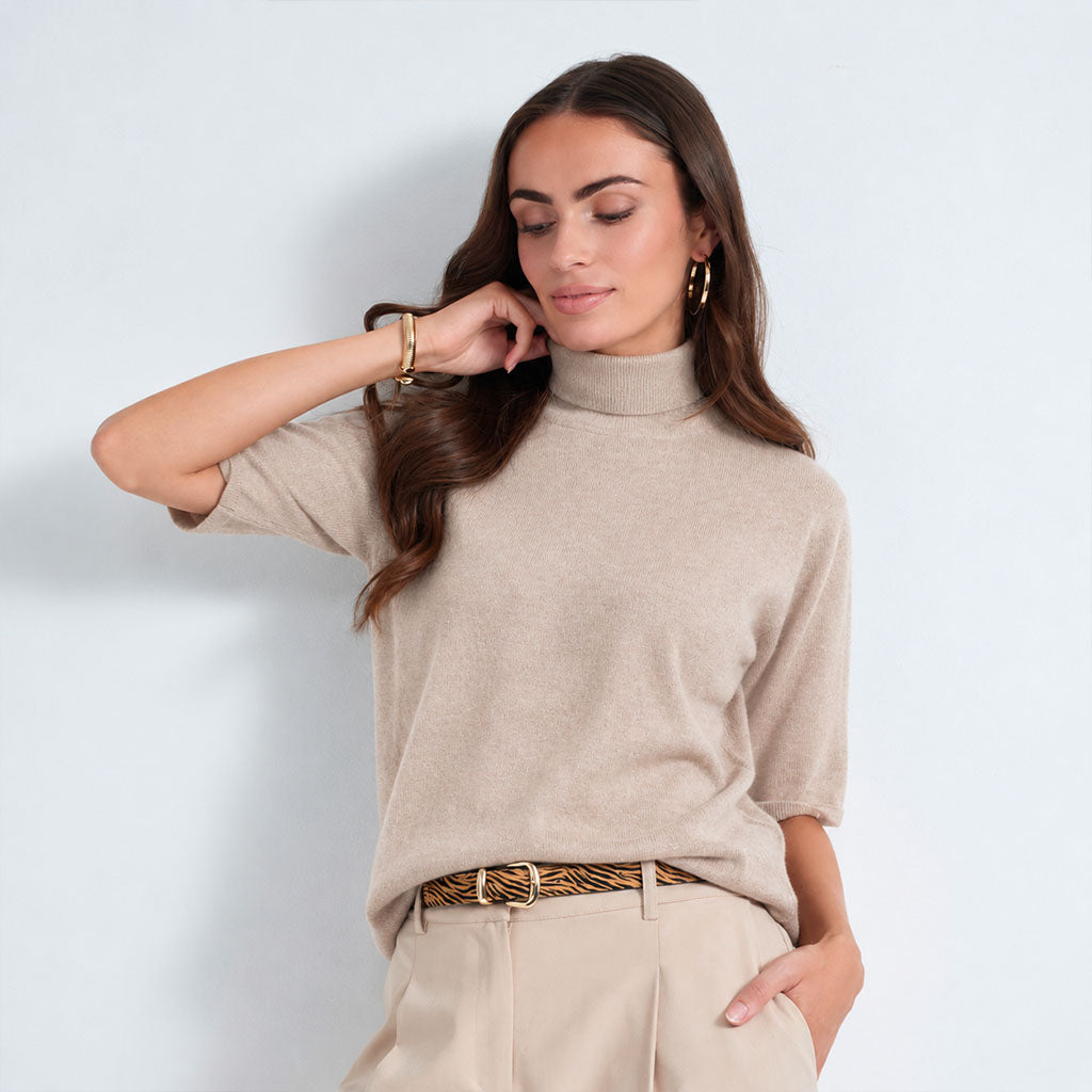 A woman wearing a beige short-sleeve mock neck knit top and beige trousers with a patterned belt, posing against a light gray studio background.