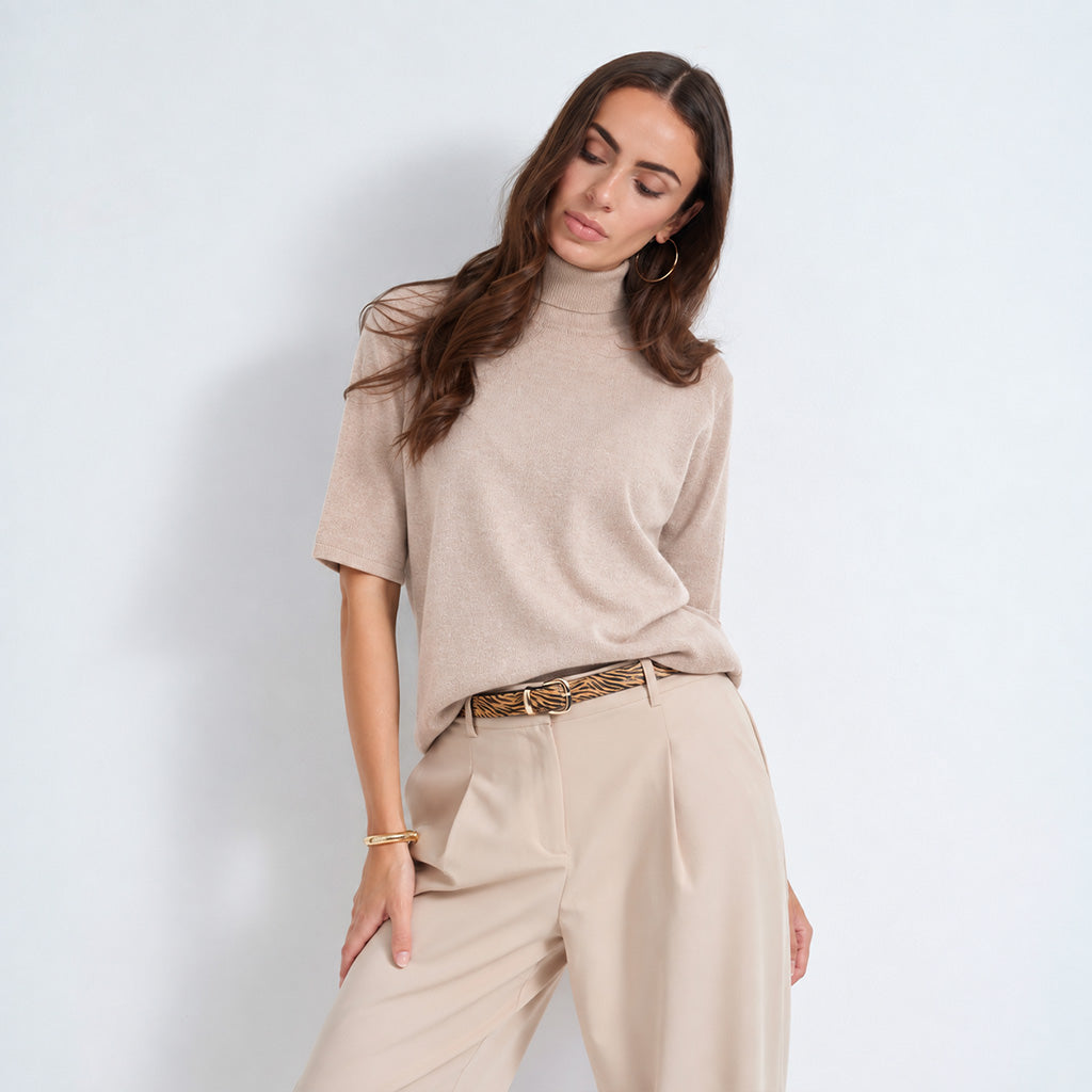 A woman modeling a beige short-sleeve turtleneck sweater paired with beige trousers and a patterned belt, set against a plain light gray studio background.