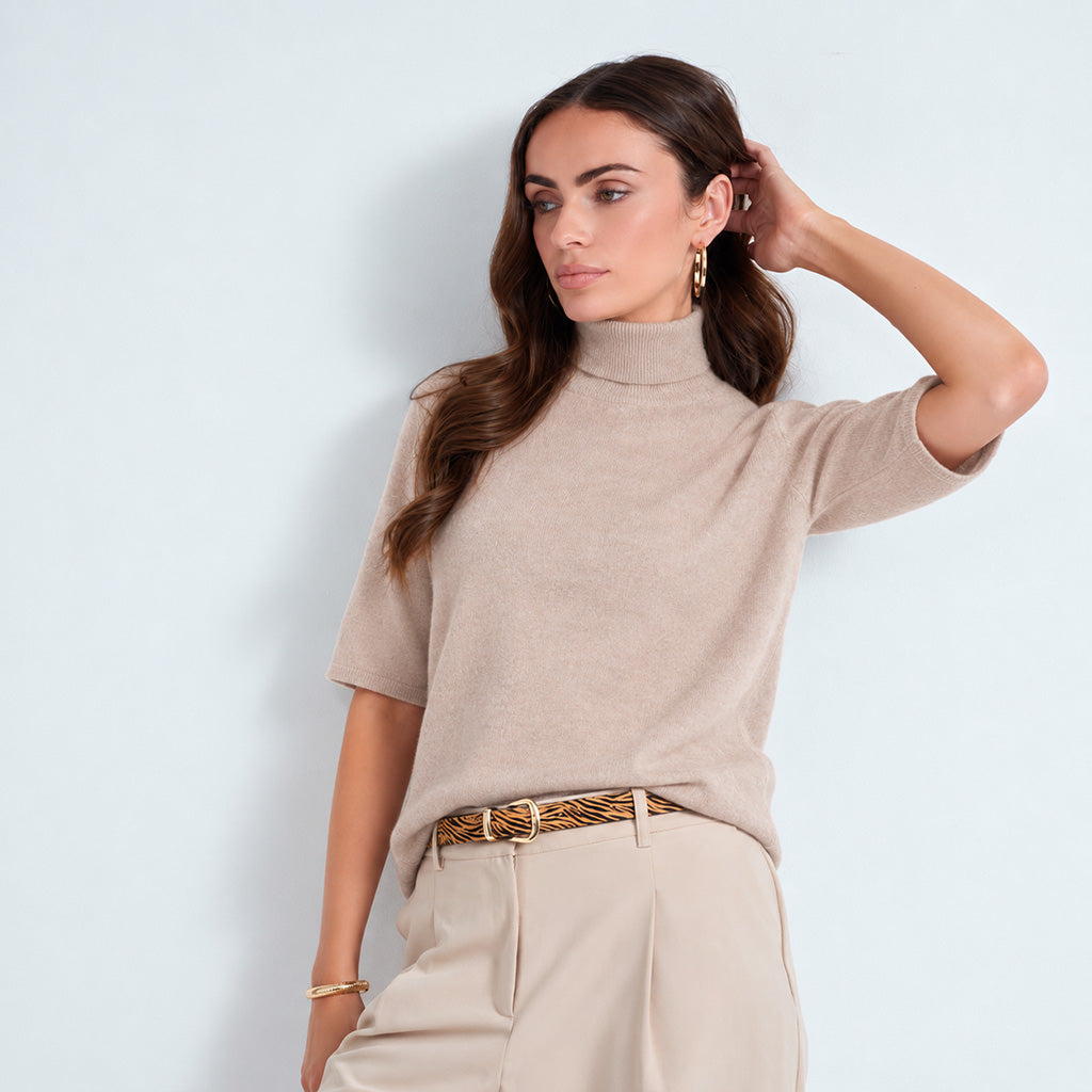 A woman wearing a beige short-sleeve mock neck knit top and cream trousers, accessorized with a patterned belt and gold jewelry, posing against a light blue wall.