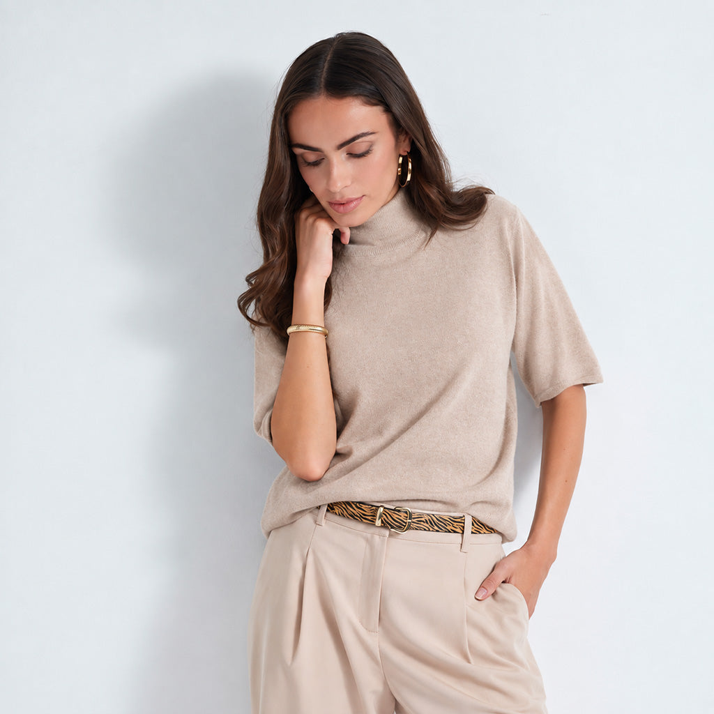 A woman wearing a beige short-sleeve mock neck knit top and matching beige trousers, styled with a patterned belt, against a light gray studio background.
