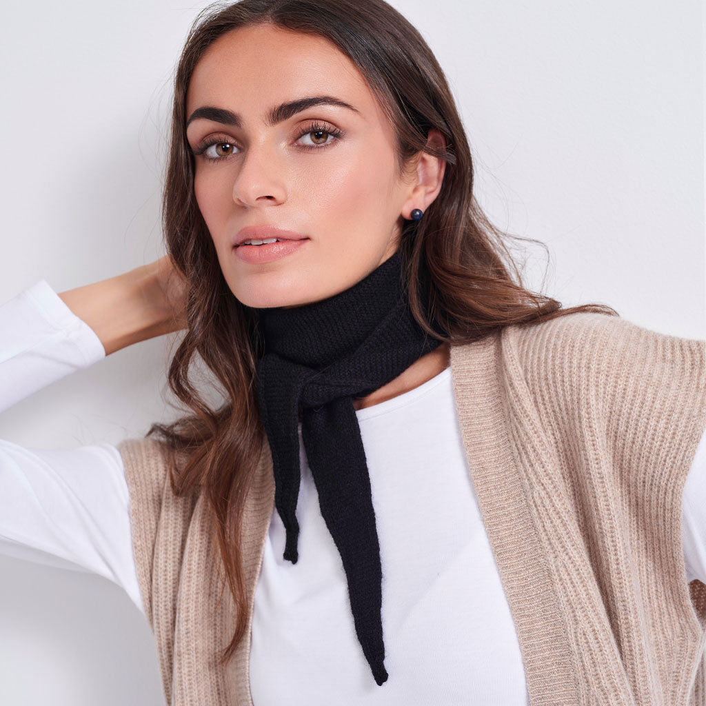 A woman wearing a black ribbed knit neck scarf tied at the front, styled over a white top and a beige ribbed knit vest against a plain light background.