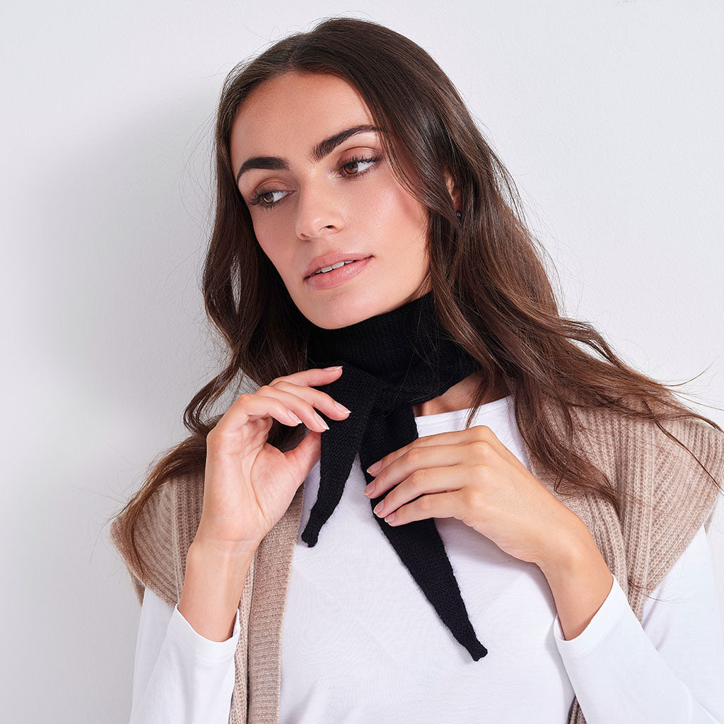 A woman wearing a black knit neck scarf tied in a bow, styled over a white top and a beige ribbed vest, set against a plain light gray background.