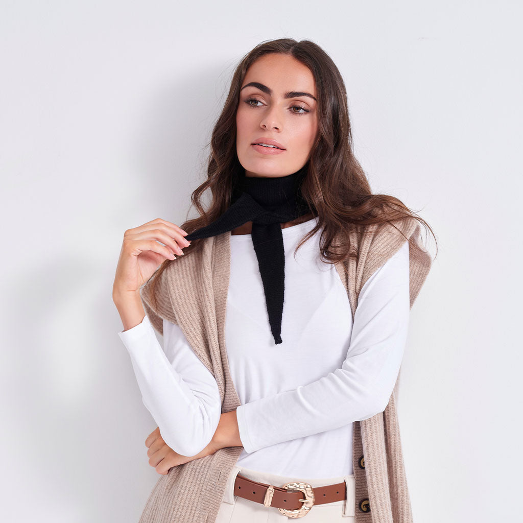 A woman modeling a beige ribbed knit sleeveless vest over a white long-sleeve top, accessorized with a black neck scarf, beige pants, and a brown belt, against a plain white studio background.