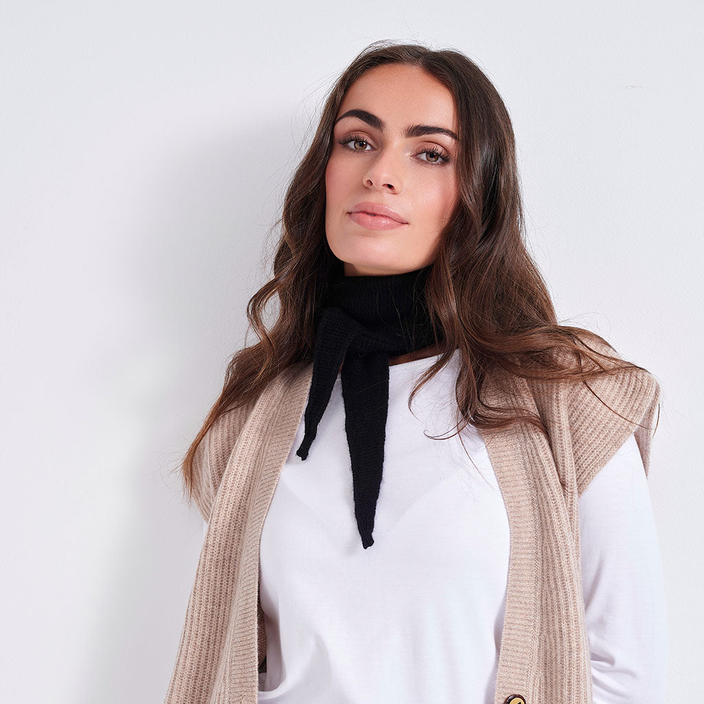 A woman with long brown hair is shown from the chest up against a plain light background, wearing a white top, a black neck scarf, and a beige ribbed sleeveless cardigan.