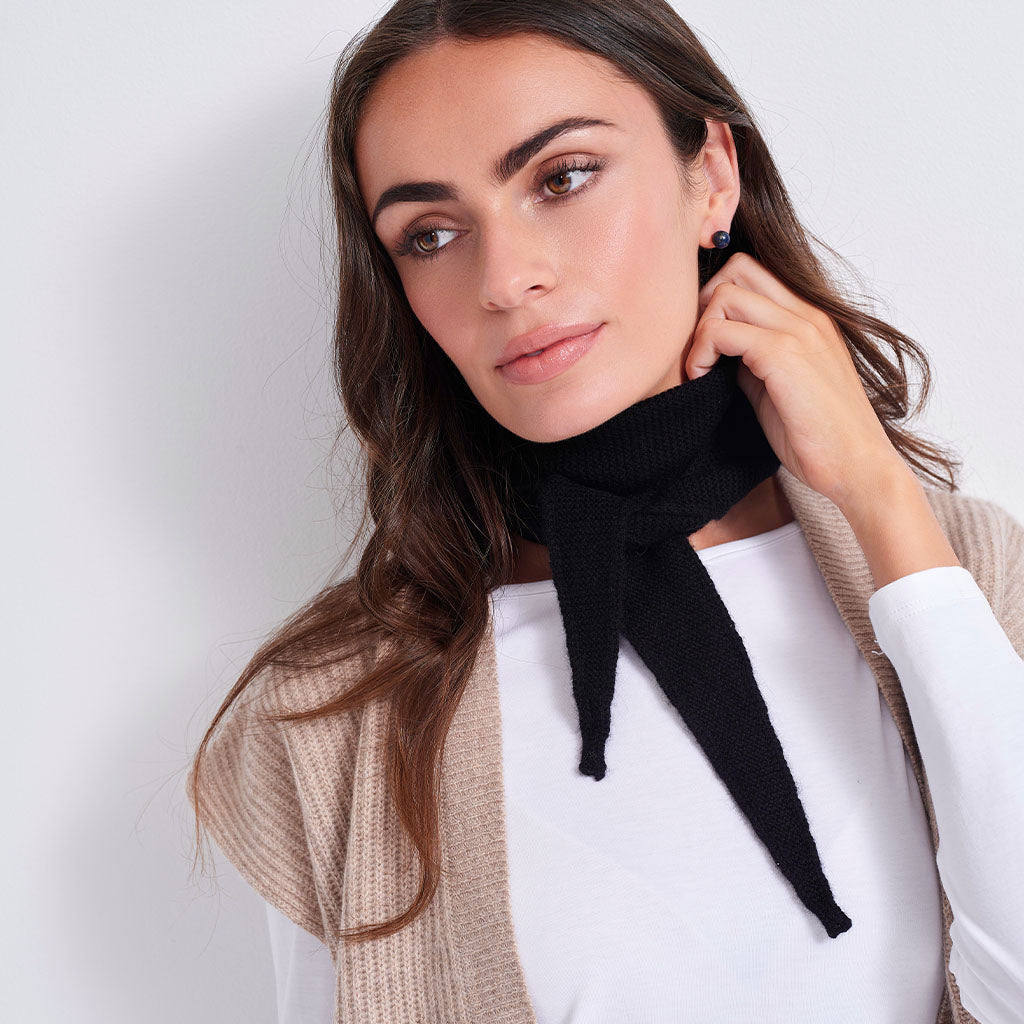 A woman wearing a black knit neck gaiter tied in a bow, styled over a white top and a beige cardigan, against a plain light gray background.