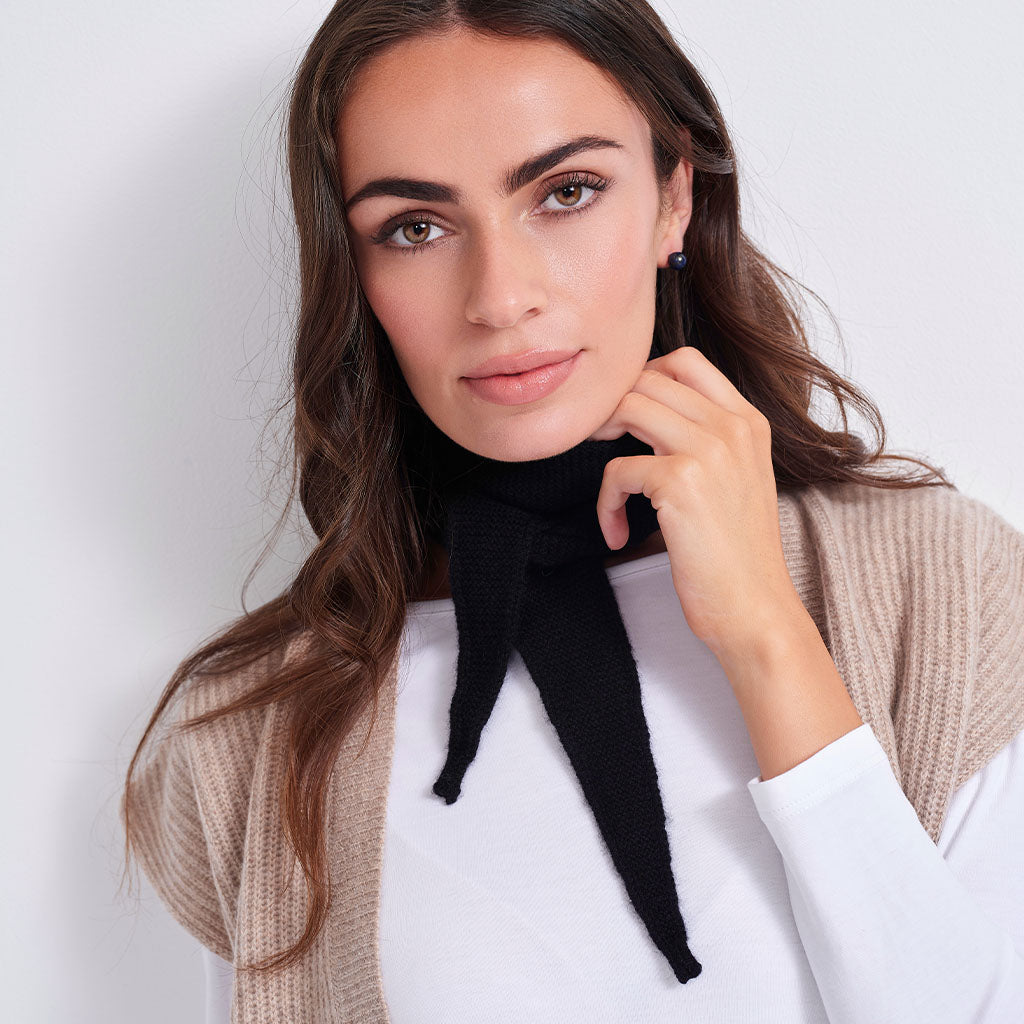 A woman wearing a black knit neck gaiter tied in a bow, styled over a white top and a beige cardigan, against a plain light gray background.