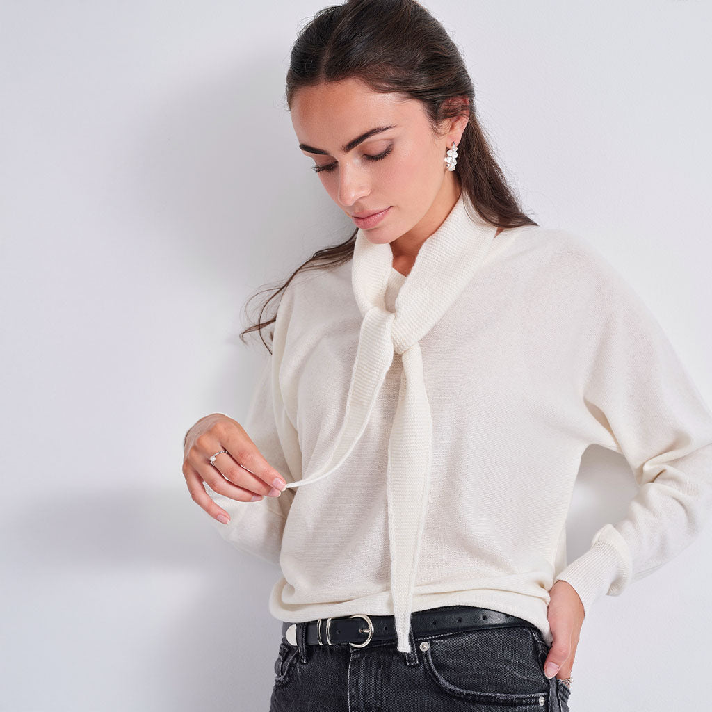 A woman wearing a cream knit neck gaiter tied in a bow, styled over a white top and a beige cardigan, against a plain light gray background.