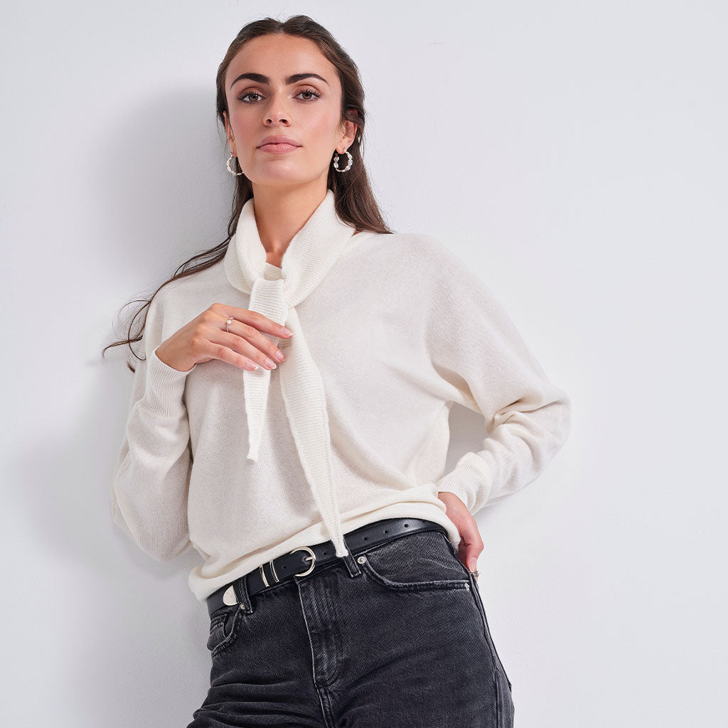 A woman modeling a cream-colored knit sweater with a wide, soft tie-neck collar, paired with black jeans and silver hoop earrings, against a plain light gray studio background.