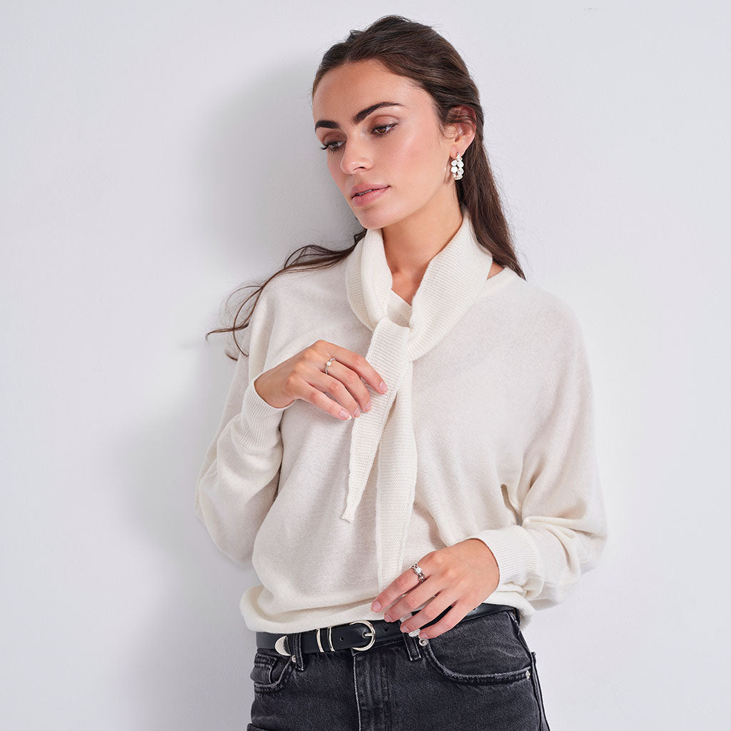 A woman wearing a cream-colored knit sweater with a wide neck and a matching knotted scarf, paired with dark gray jeans, posing against a light gray wall.