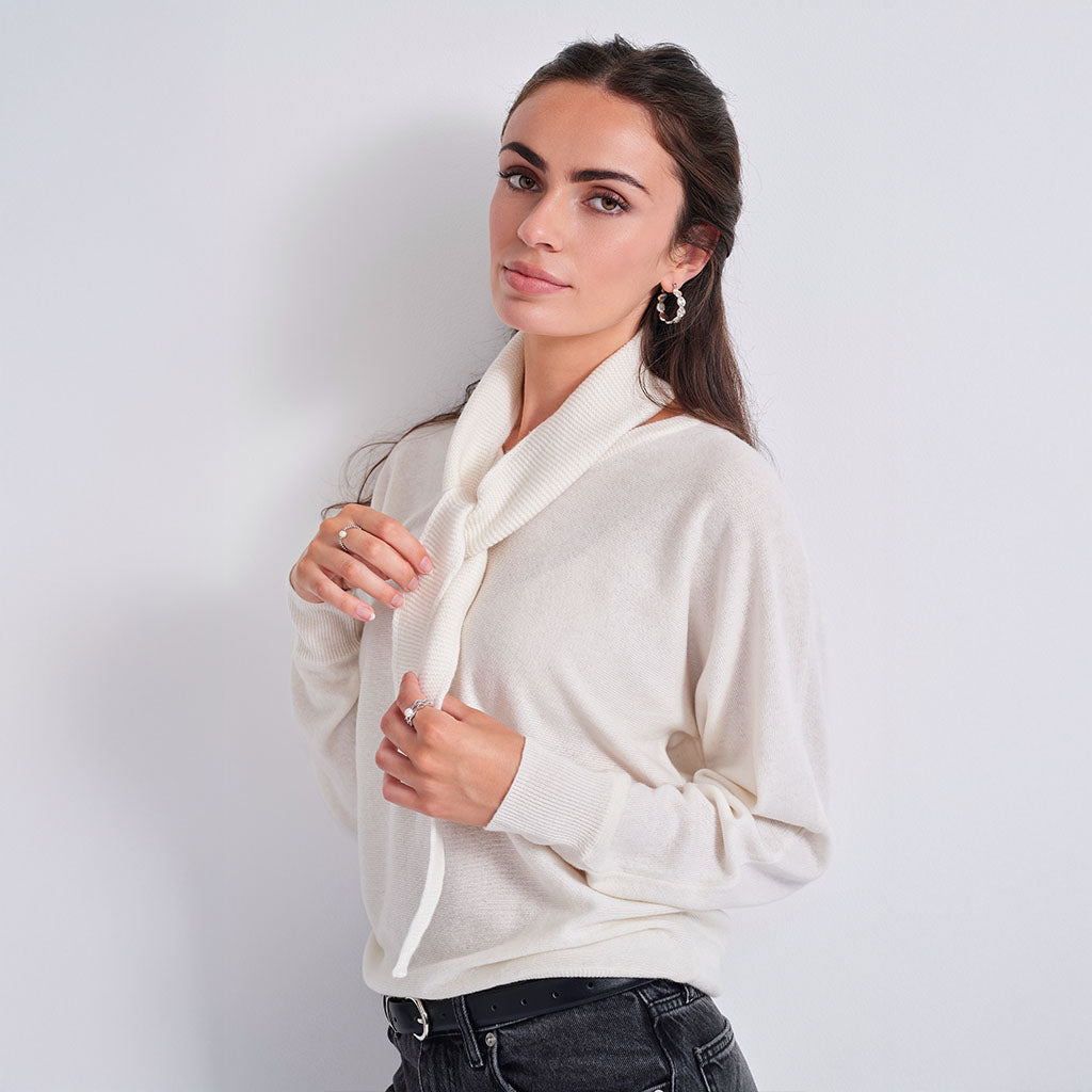 A woman wearing a cream-colored knit sweater with a wide tie-neck detail, paired with dark jeans, posing against a light gray studio background.