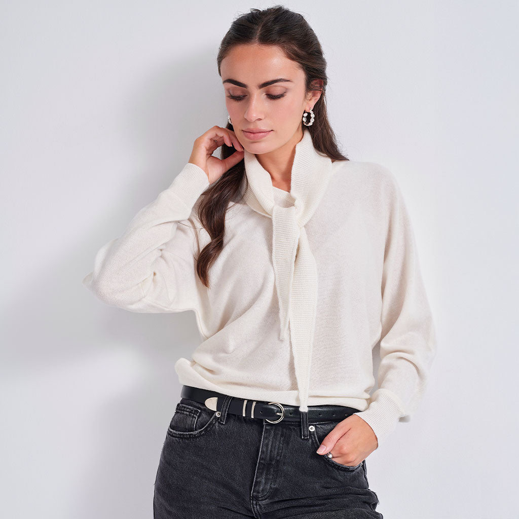 A woman wearing a cream-colored knit sweater with a wide cowl neck and a front tie detail, paired with black jeans and a black belt, posing against a light gray studio background.