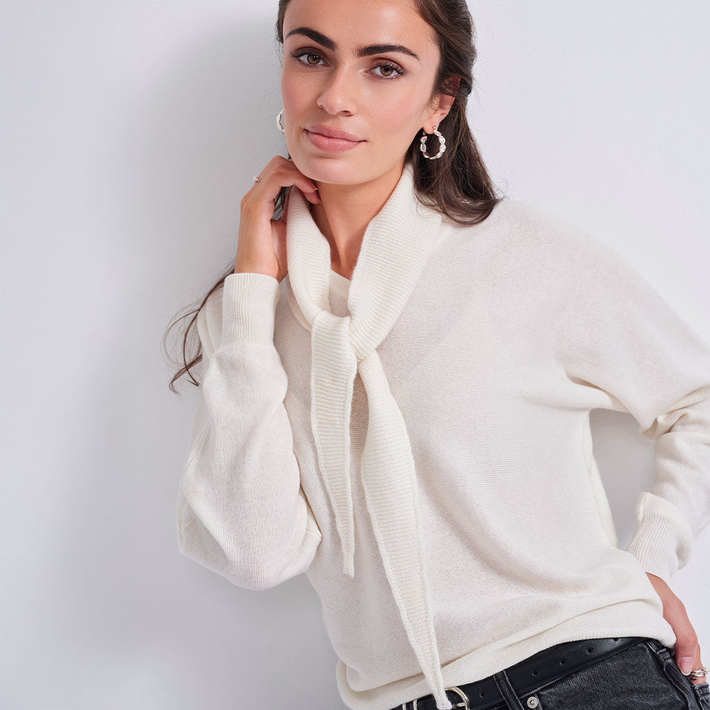 A woman wearing a cream-colored knit sweater with a wide ribbed collar and a matching tie-neck detail, paired with dark jeans and silver hoop earrings, set against a plain light gray background.