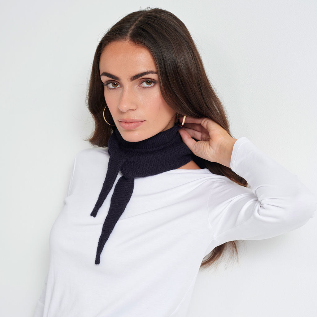 A woman wearing a dark navy blue knit turtleneck scarf with a tie detail, styled over a white long-sleeve top against a light gray background.