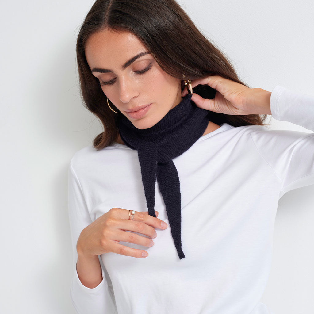A woman wearing a dark navy blue knit turtleneck scarf with a tie detail, styled over a white long-sleeve top against a light gray background.