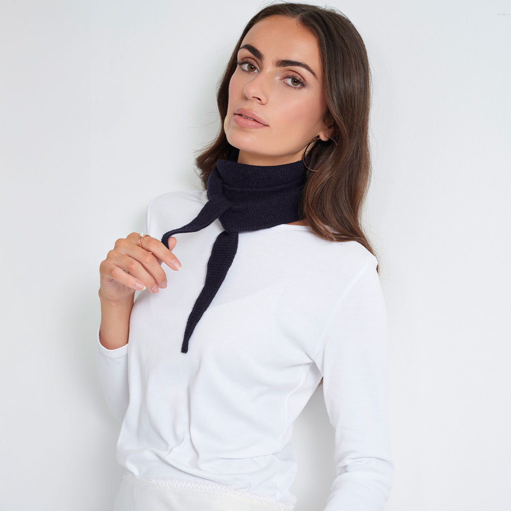 A woman wearing a white long-sleeve top and a dark navy knit neck gaiter tied around her neck, photographed against a clean white studio background.