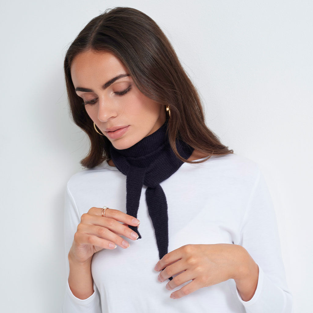 A woman wearing a white long-sleeve top and a black knit turtleneck scarf with a tie detail, posed against a light gray studio background.