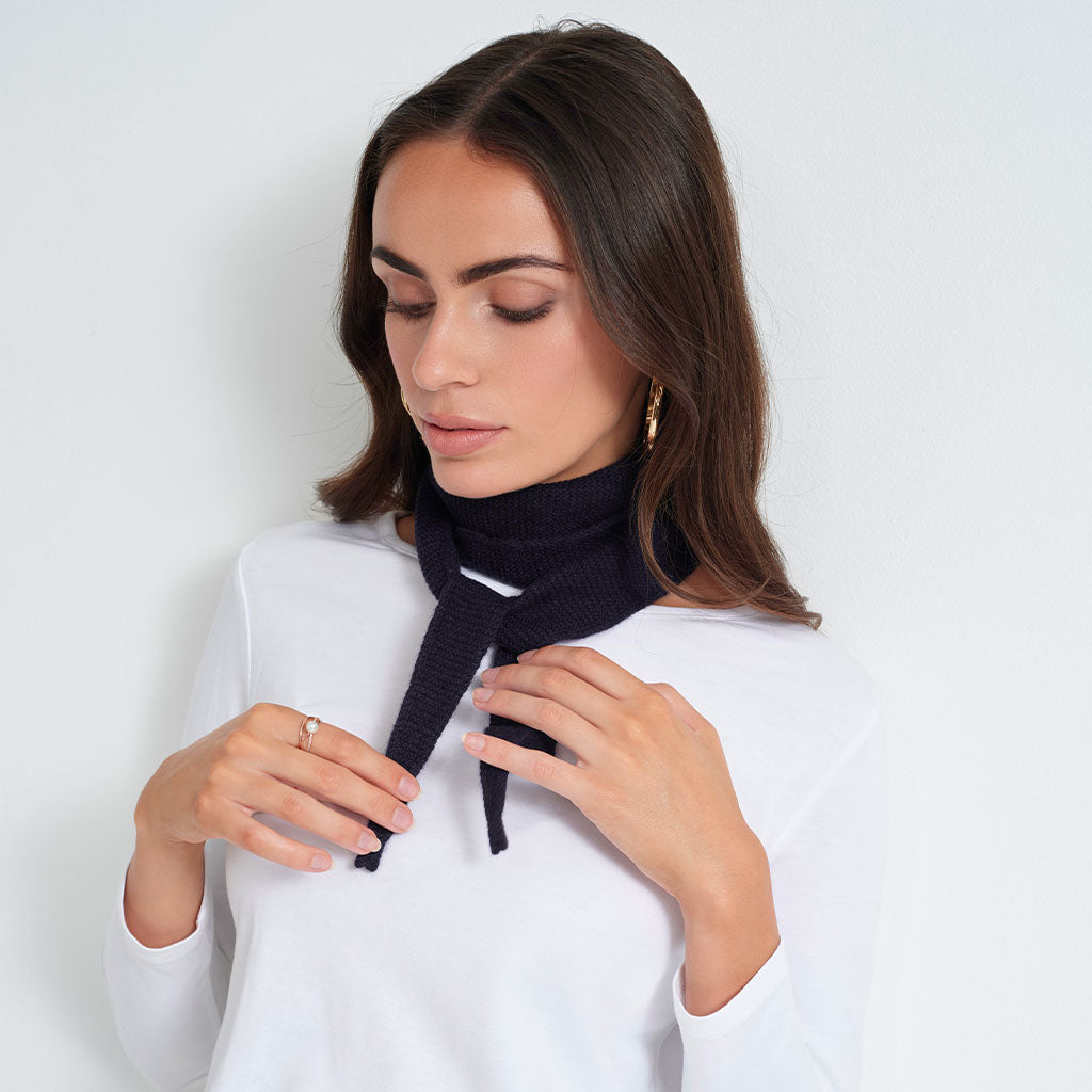 A woman wearing a white long-sleeve top and a black ribbed knit neck warmer scarf, adjusting it around her neck against a plain white background.