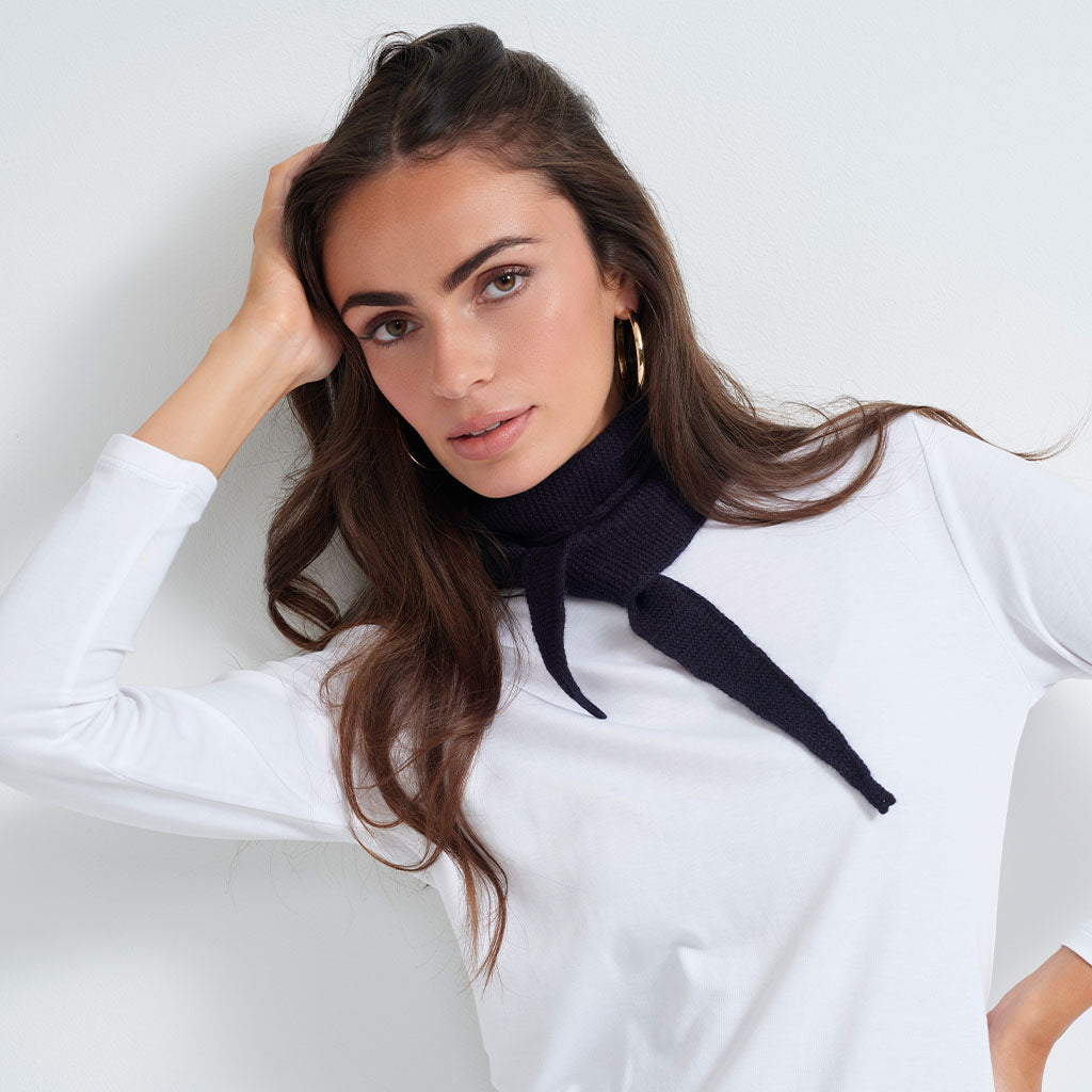 A woman wearing a white long-sleeve top and a black knit neck scarf, posing against a clean white background.