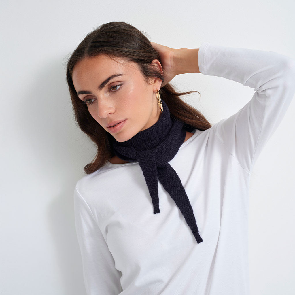 A woman wearing a dark navy blue ribbed knit neck warmer tied at the front, styled over a white long-sleeve top against a plain white background.