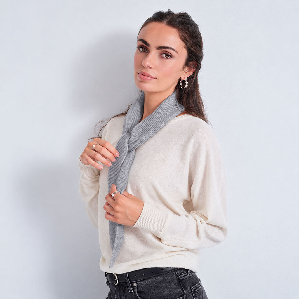A woman wearing a cream-colored knit sweater and a gray striped knit scarf, posing against a light gray studio background.