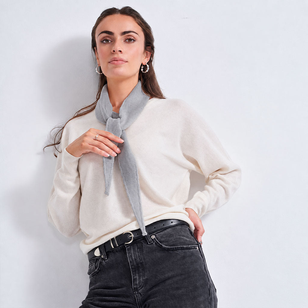 A woman modeling a light gray knit scarf tied around her neck, worn over an off-white sweater and black jeans against a plain light gray wall.