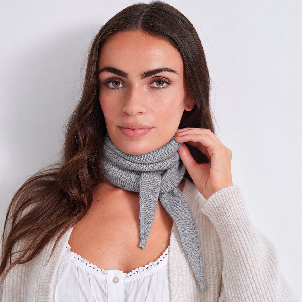 A woman with long brown hair models a light gray knit scarf tied around her neck, wearing a white lace-trimmed top and a cream-colored cardigan against a plain light gray background.