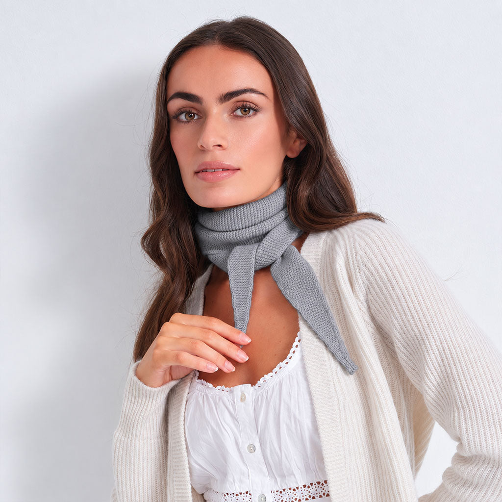 A woman wearing a light gray knit cowl scarf tied around her neck, styled with a white lace-trimmed top and a cream ribbed cardigan, set against a plain light gray background.
