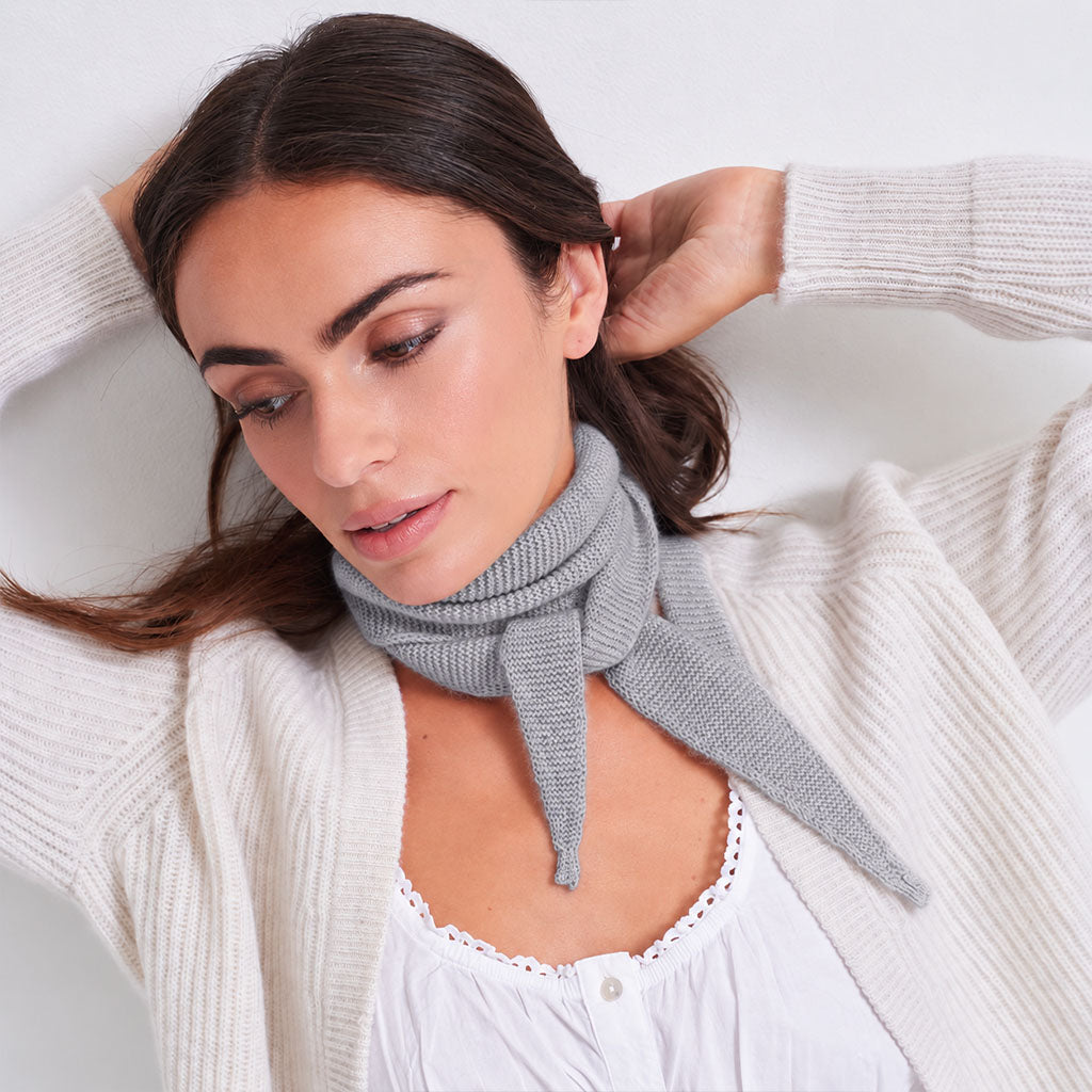 A woman wearing a light gray knitted cowl scarf tied around her neck, styled with a white lace-trimmed top and a cream ribbed cardigan against a plain light background.
