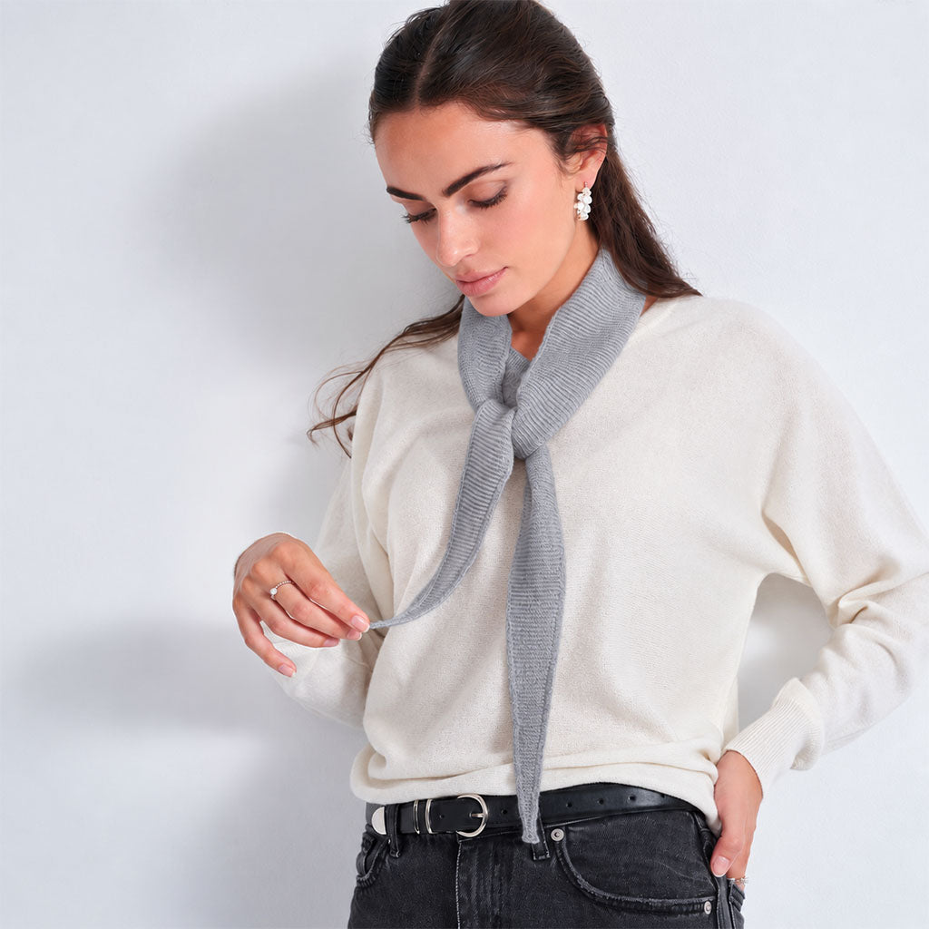 A woman wearing a cream-colored knit sweater and a light gray knitted tie-neck scarf, paired with black jeans and a black belt, set against a plain light gray studio background.
