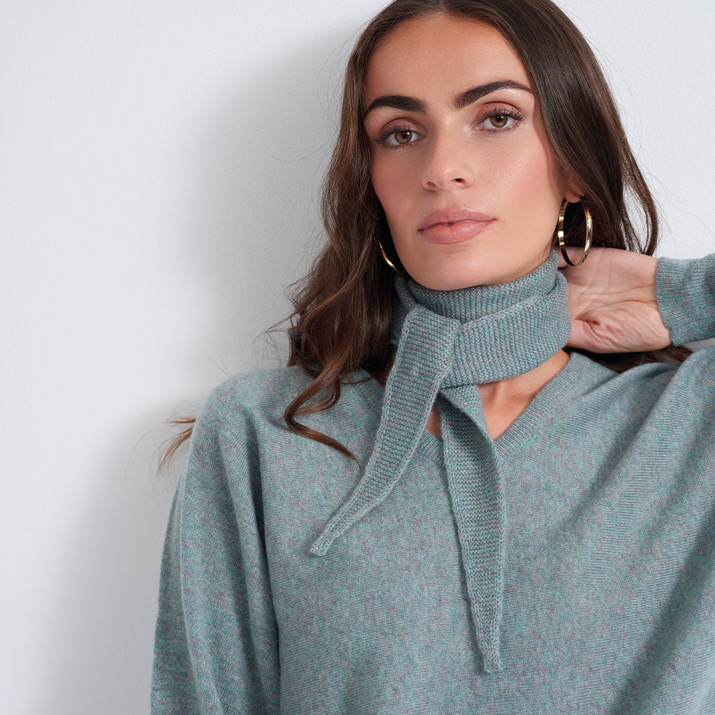 A woman wearing a light blue-gray knit sweater with a high ribbed turtleneck collar tied at the front, styled with large gold hoop earrings against a plain light gray background.