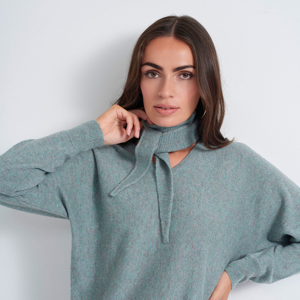 A woman wearing a sage green knit sweater with a tie-neck detail, posing against a light gray studio background.