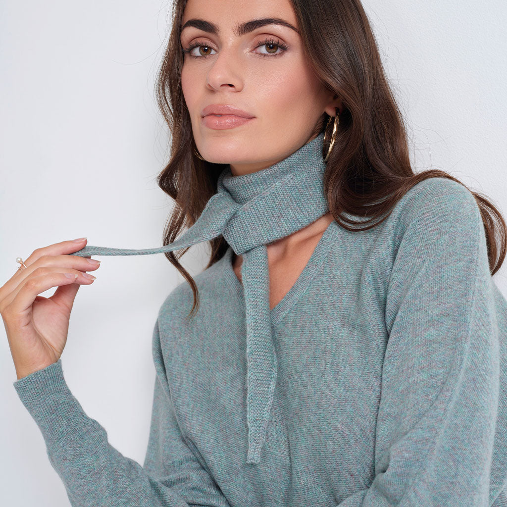 A woman wearing a heathered sage green knit cowl neck sweater and matching knit scarf, posing against a light gray background.