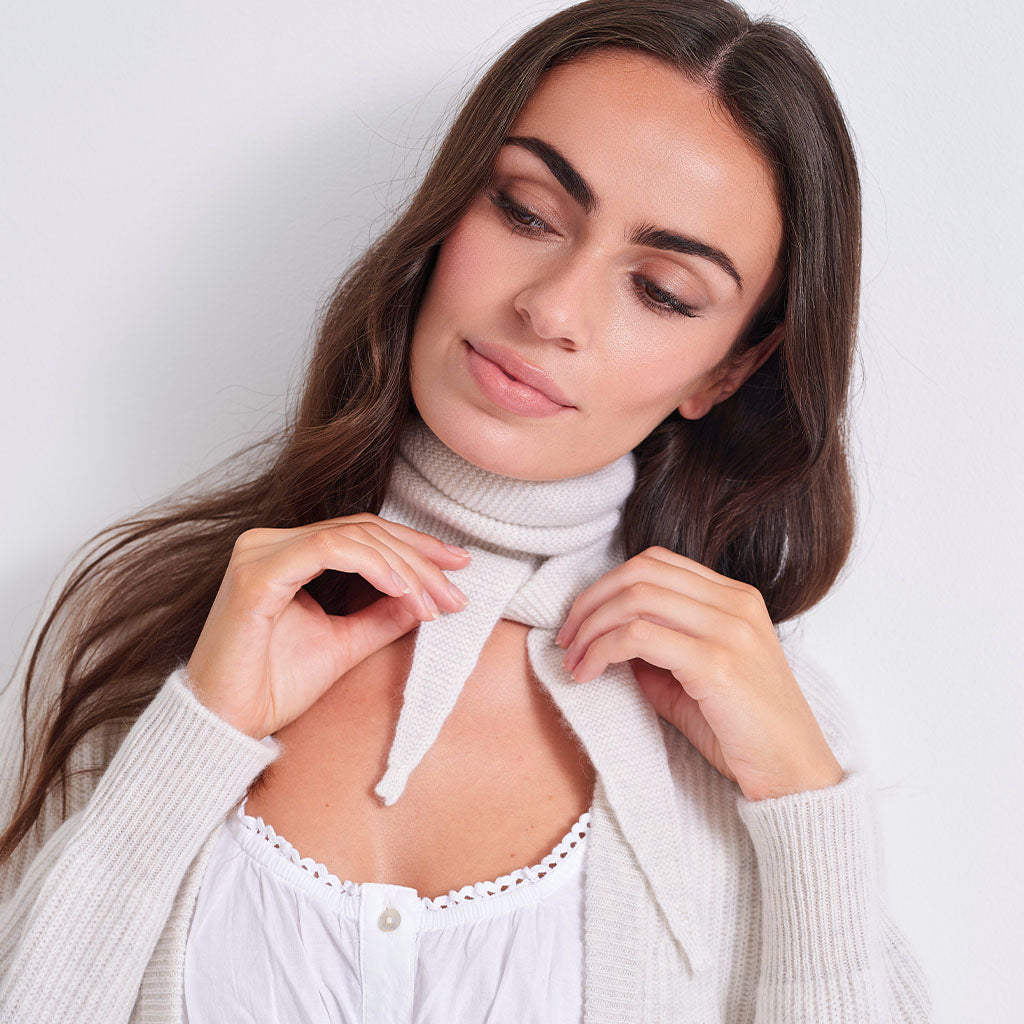 A woman with long dark brown hair wearing a cream-colored ribbed knit turtleneck sweater tied at the neck over a white lace-trimmed top, set against a plain light gray background.