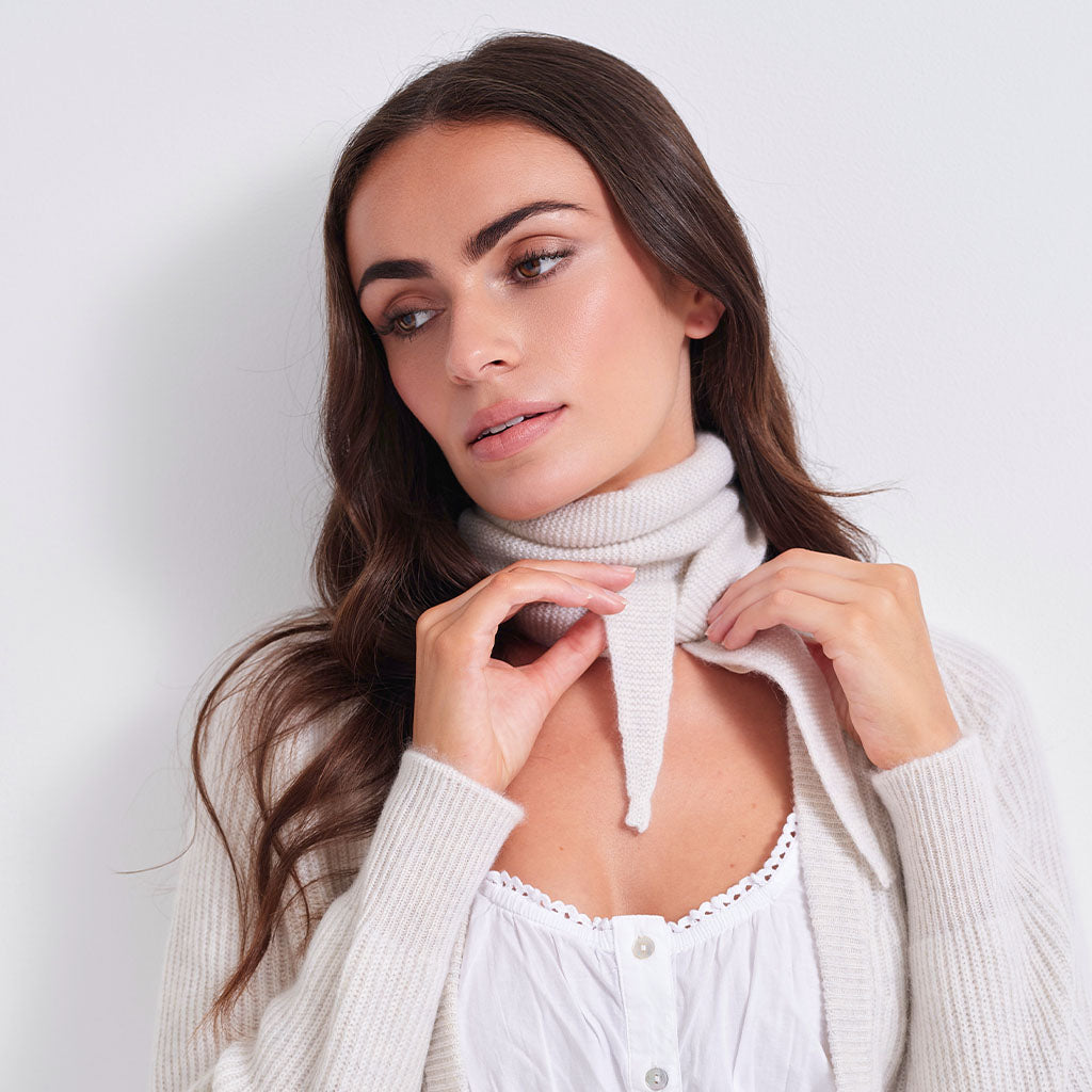 A woman with long brown hair models a cream-colored ribbed knit cowl neck scarf tied under her collar over a white top, set against a plain light gray background.
