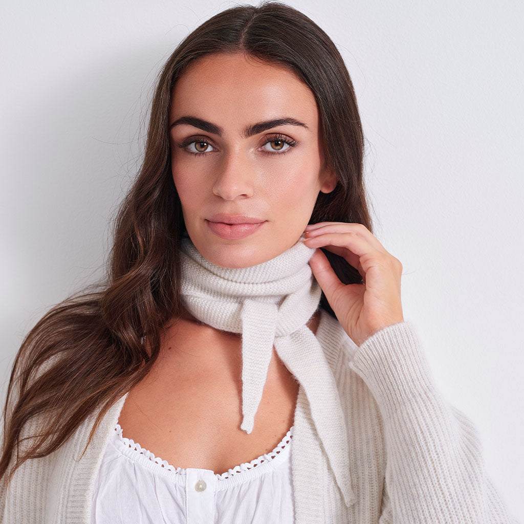 A woman with long brown hair models a cream-colored ribbed knit cowl scarf tied around her neck, paired with a white lace-trimmed top and a white cardigan against a plain light gray background.