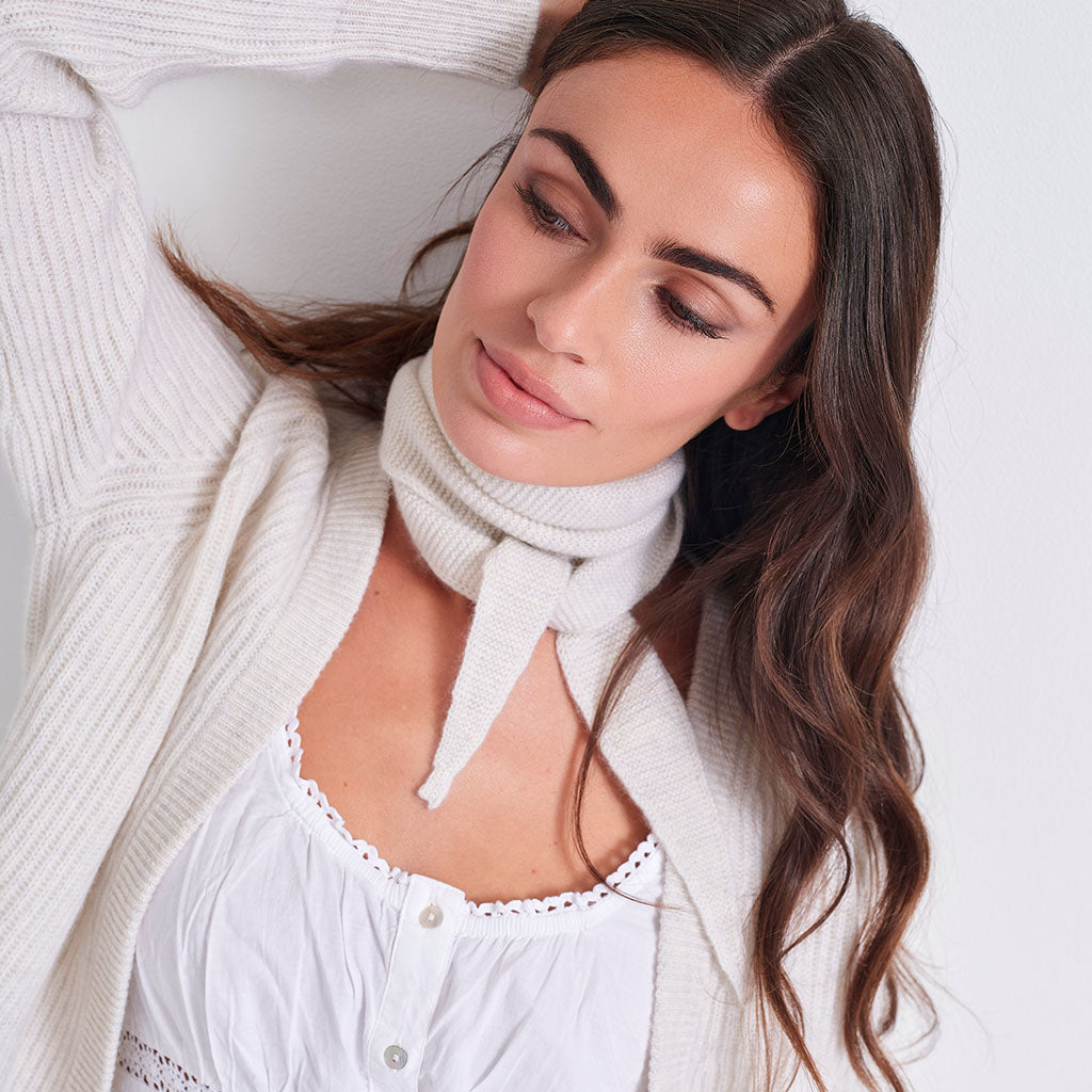 A woman wearing a white ribbed knit cowl neck scarf tied at the front, layered over a white camisole and a cream-colored cardigan, set against a plain white background.