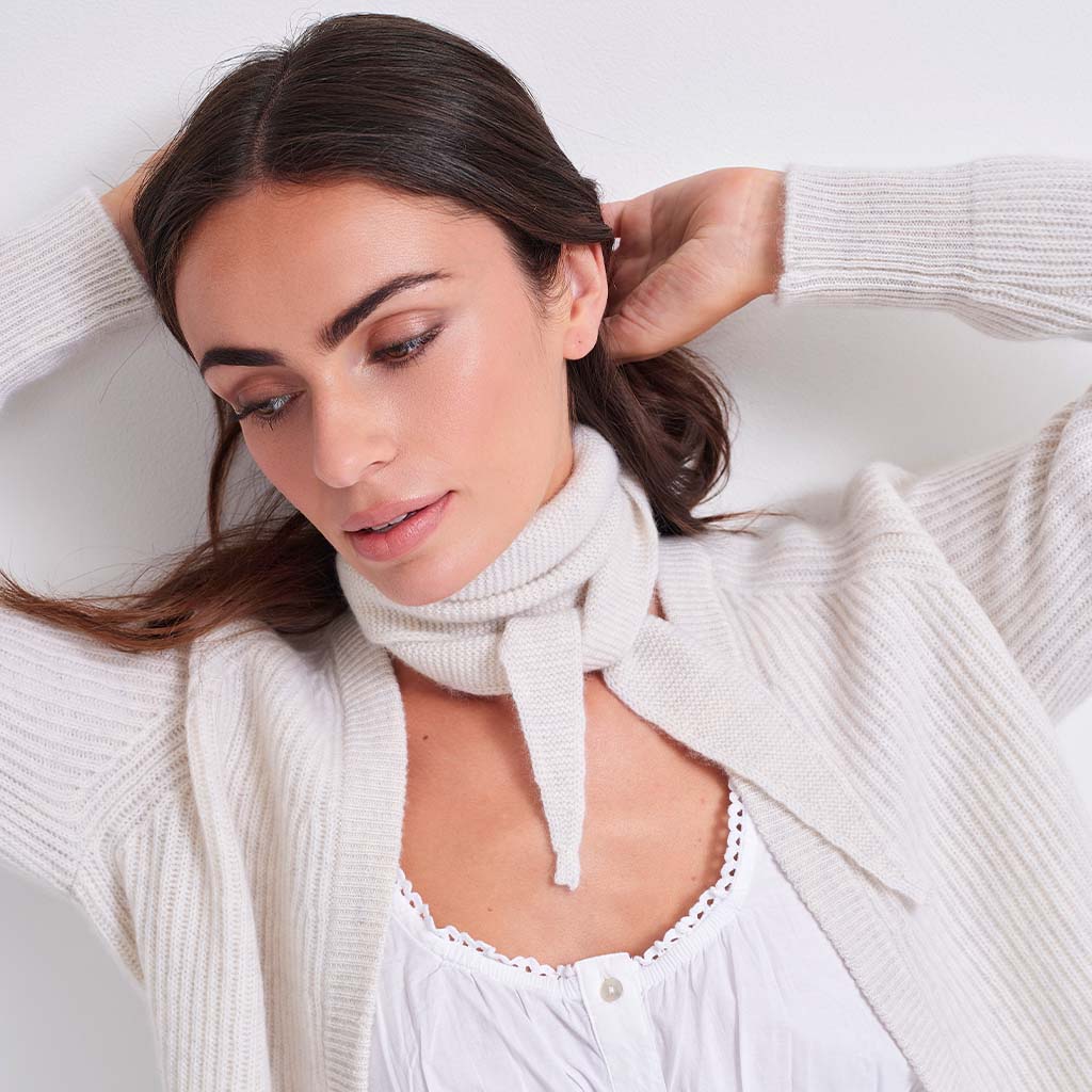 A woman wearing a cream-colored ribbed knit cardigan sweater over a white top, with a matching cream knit scarf tied around her neck, set against a plain light background.