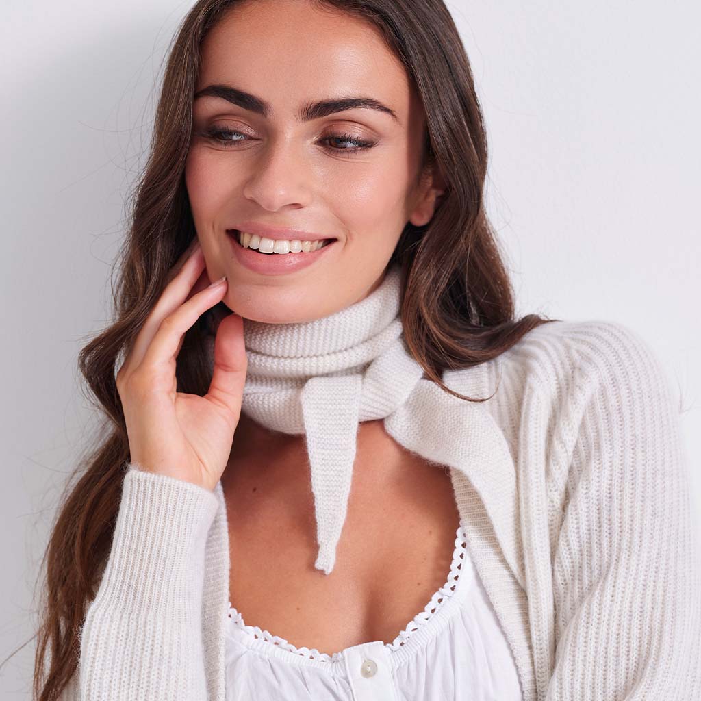 A woman with long brown hair wearing a white ribbed knit turtleneck scarf tied at the neck, layered over a white top and a cream-colored cardigan, against a plain light background.