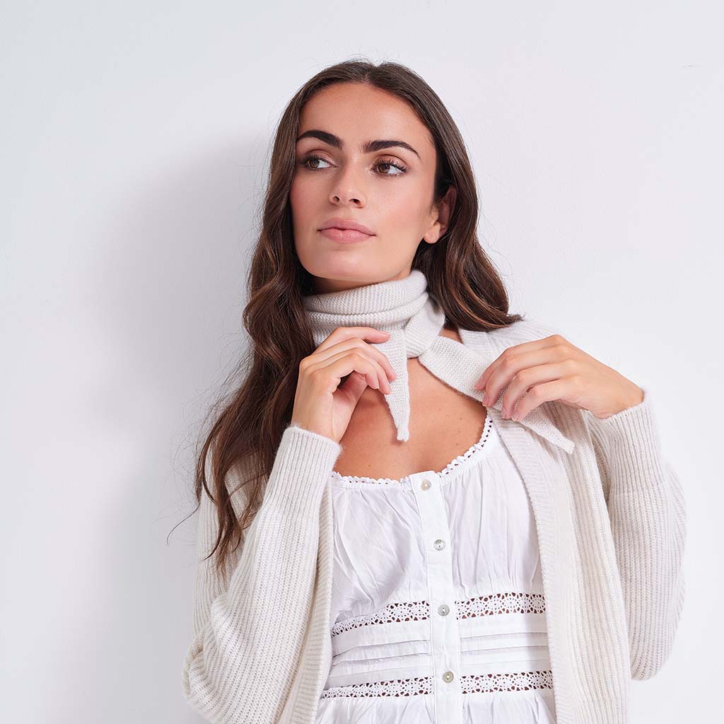 A woman with long brown hair wearing a white eyelet top, a cream knit cardigan, and a cream knit neck warmer, posing against a plain white background.