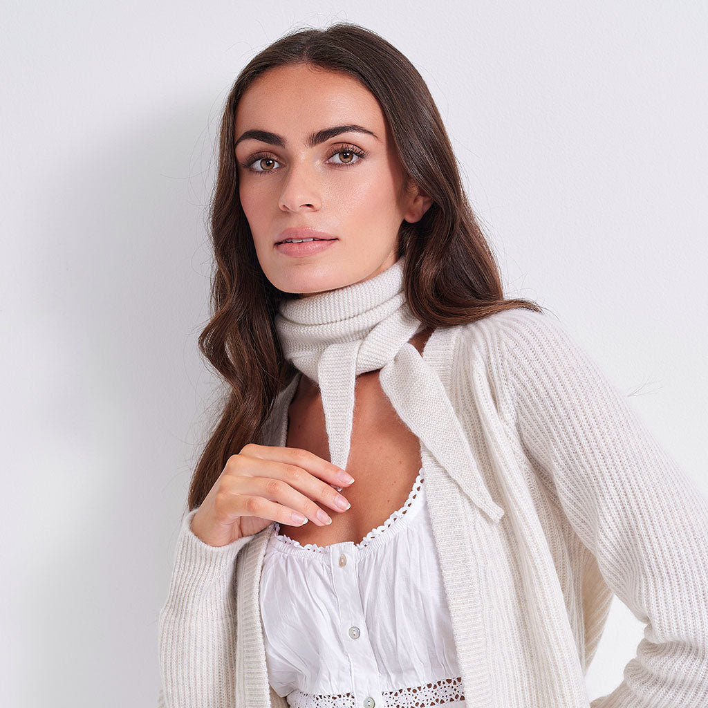 A woman wearing a cream ribbed knit cardigan over a white top, with a matching cream knit cowl scarf tied at the neck, set against a plain light gray background.