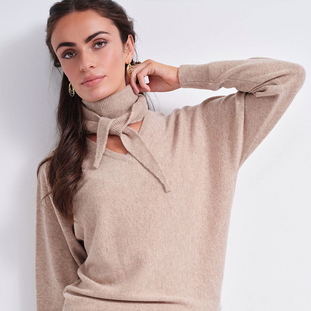 A woman wearing a beige knit turtleneck sweater with a tie detail at the neck, posed against a plain light gray background.
