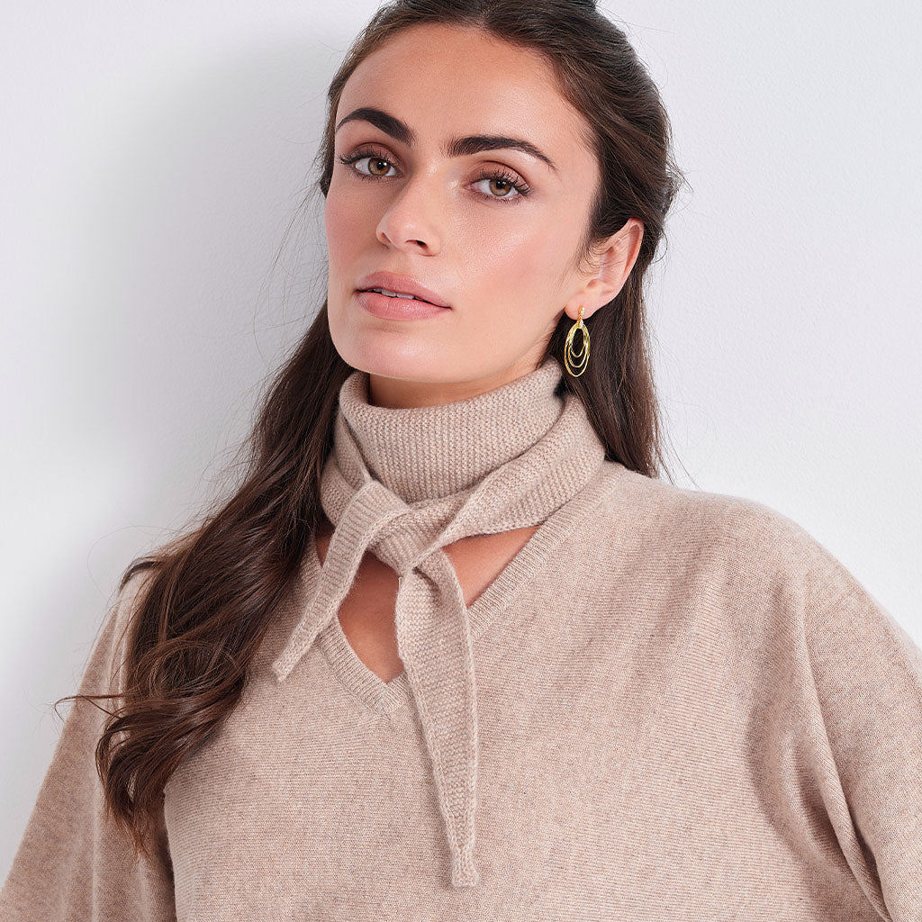 A woman wearing a beige knit sweater with a high folded collar and a front tie detail, paired with gold hoop earrings, against a plain light gray background.