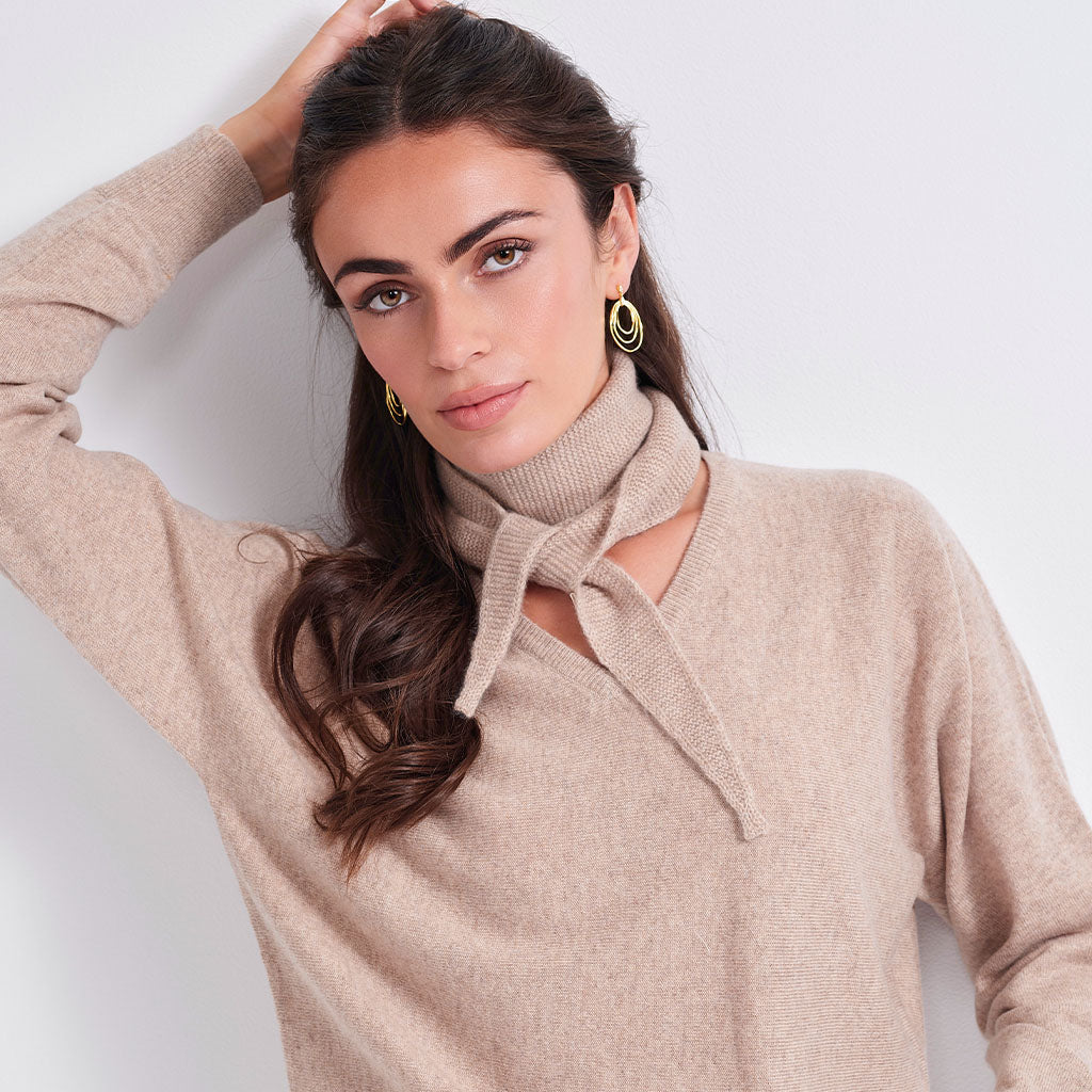 A woman wearing a beige knit sweater with a tie-neck detail, styled with gold hoop earrings against a plain light gray background.